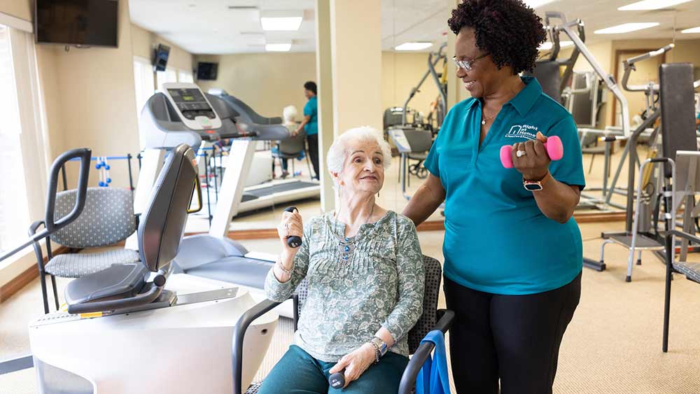 senior-woman-seating-exercising-with-standing-caregiver-in-workout-room