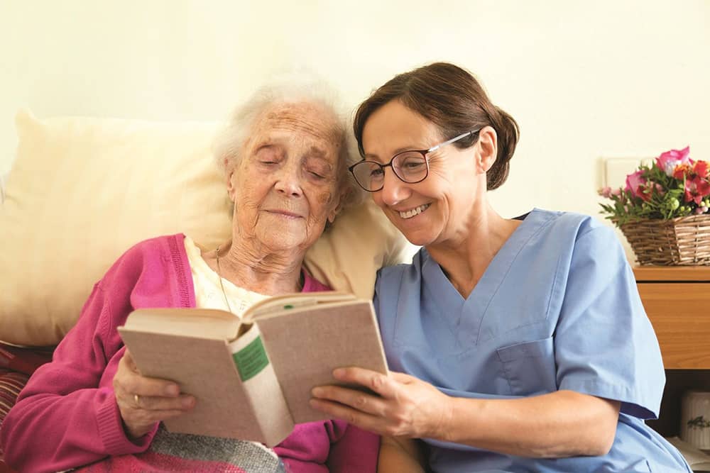 Right at Home Caregiver Seated on Bed Reading Book with Elderly Woman