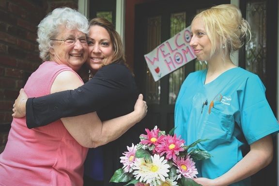 Senior Woman Receiving Welcome Home Hug while Right at Home Female Caregiver in Blue Scrubs Holding Flowers Watches