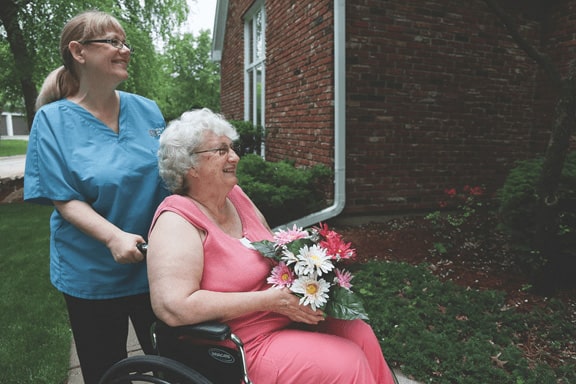 Caregiver in Blue Scrubs Walking with Elderly Woman in Wheelchair Holding Flowers