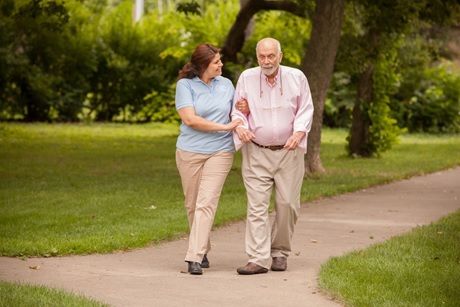Female Right at Home caregiver in blue Right at Home shirt walking outside on tree lined sidewalk path with elderly gentleman wearing pink shirt