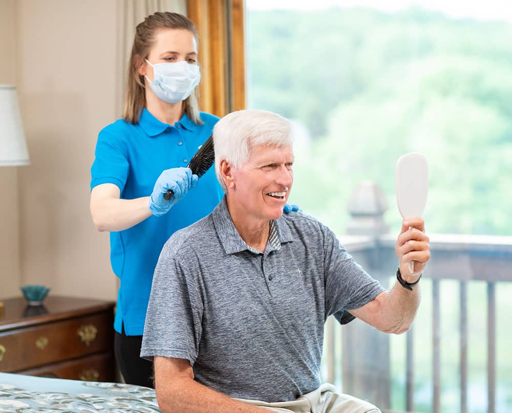 caregiver wearing PPE brushing the hair of senior adult