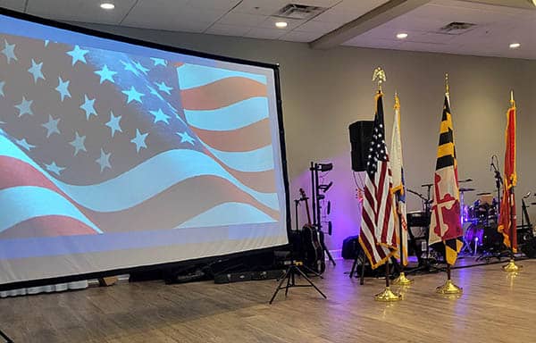 Indoor setting with an American flag projected on a screen, with the American and Maryland state flags on flagpoles to the right.