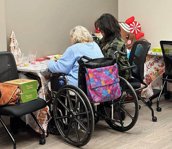 A female caregiver and elderly client sit at a table and decorate cookies together.