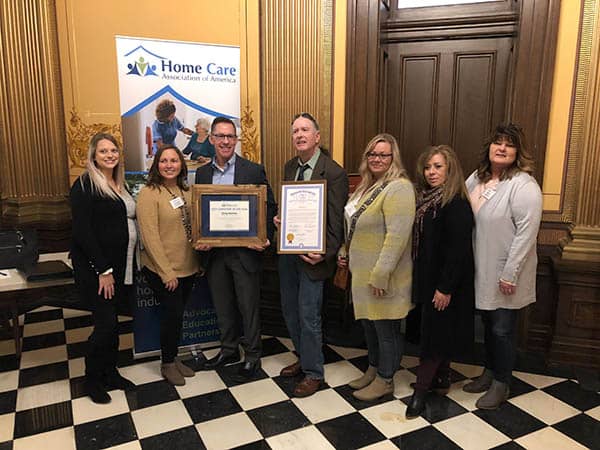 Right at Home of Genesee County's Greg Gorton holding his 2021 National Caregiver of the Year Award from the HCAOA, standing with Owner Scott Hall and other staff members of the office.