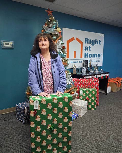 Pamela standing in front of a Christmas tree holding a large gift.