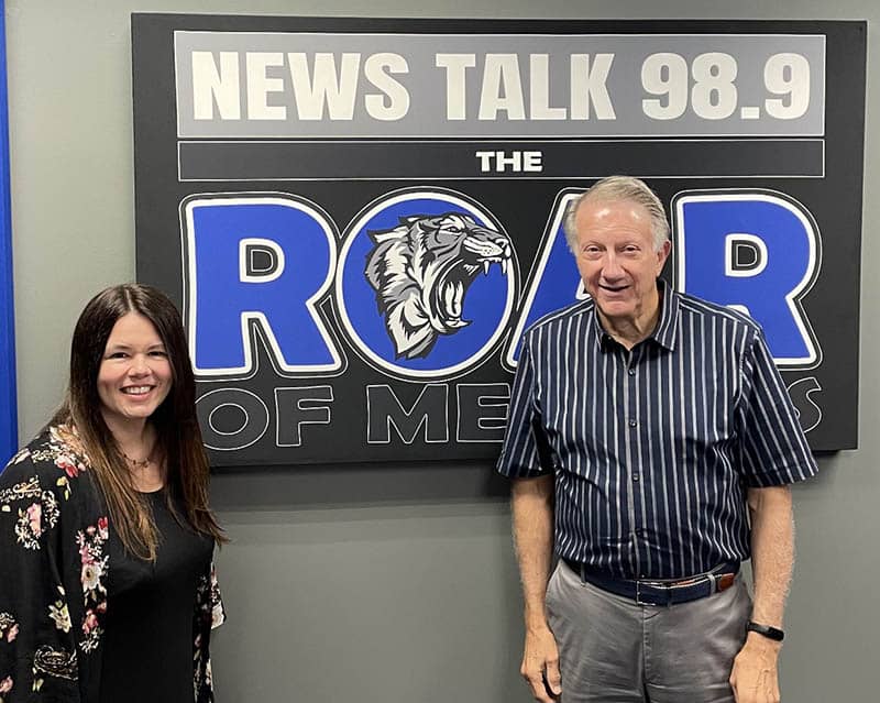 Carol standing with Jim Shoemaker in front of 98.9 The ROAR sign.