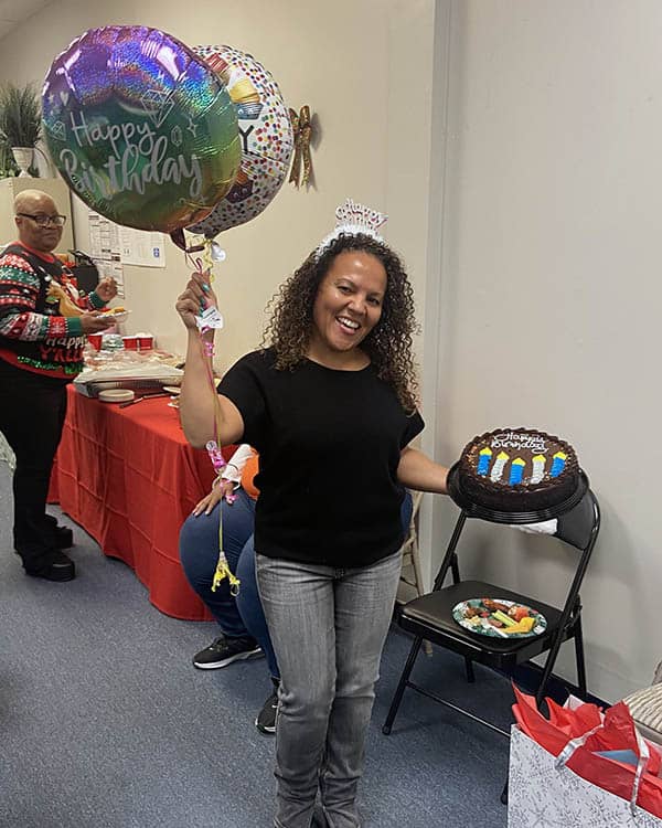 Melanie stands holding balloons in one hand and a cake in the other.