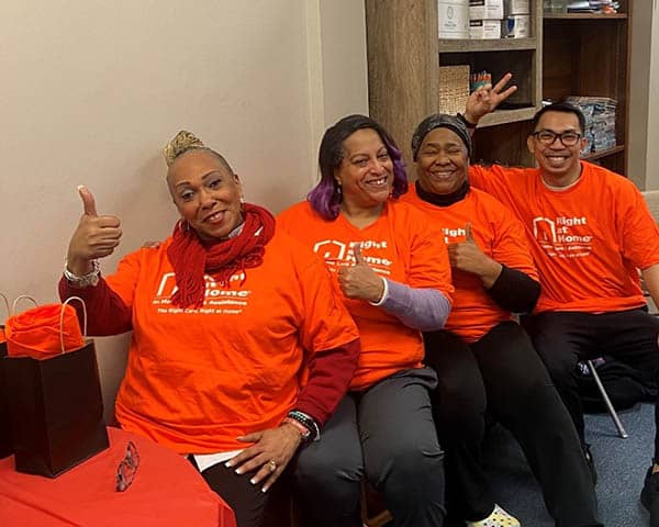 A group of four caregivers wearing bright orange Right at Home t-shirts giving a thumbs-up.