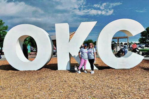 Two young women pose standing next to the OKC sculpture and sign at the Oklahoma City Parkinson's Walk Fundraiser