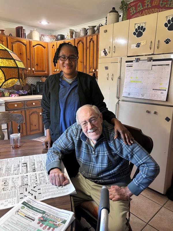 Black female caregiver standing next to sitting senior white male client