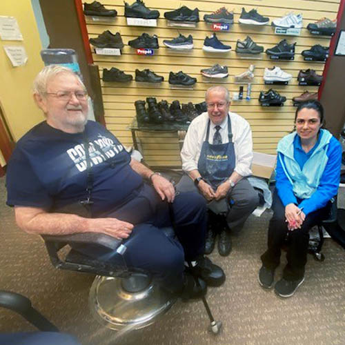 Senior Male Veteran Sitting in a Chair in a Cobbler's Shop with the Cobbler and His Female Assistant