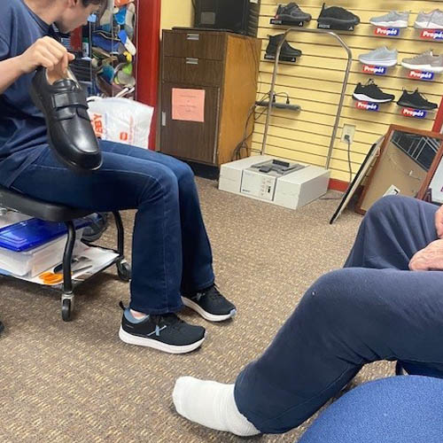 Senior Male Veteran Sitting in a Chair in a Cobbler's Shop with a Female Assistant Opening a Box of Custom Formed Shoess