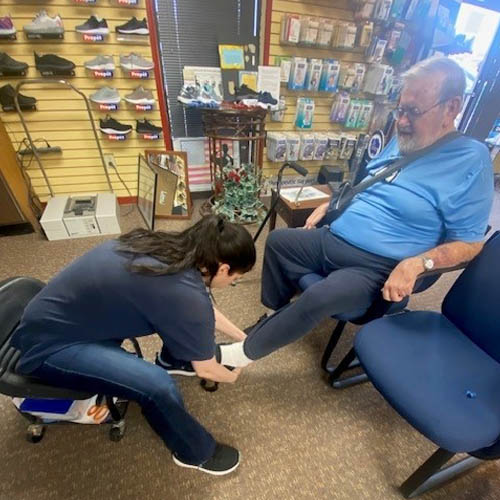 Senior Male Veteran Sitting in a Chair in a Cobbler's Shop with a Female Assistant Putting on a Pair of Custom Formed Shoes