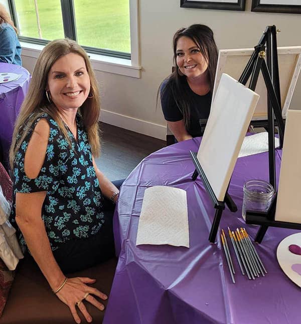 Two women smile while seated in front of a blank canvas