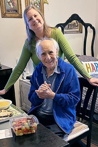 Senior Male Client Sitting at Table In Front of Birthday Treats with A Female Caregiver Standing Over His Shoulder