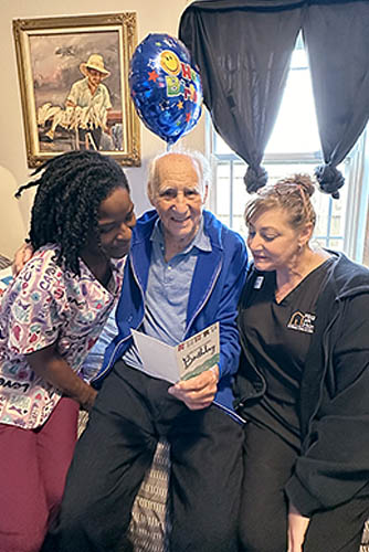 Two Female Caregivers Sitting With A Male Client Reading a Birthday Card