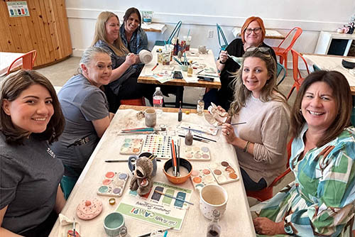 Group of men and women smiling at the camera sitting at a table working on painting pottery