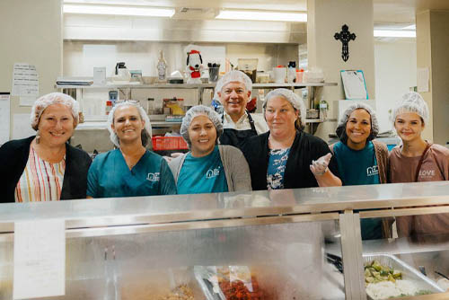 Seven Mission of Yahweh faith-based male and female shelter volunteers standing side-by-side behind a food preparation counter with gloves and hair nets and smiling at the camera