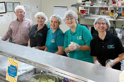 Five Mission of Yahweh faith-based male and female shelter volunteers standing side-by-side behind a food preparation counter with gloves and hair nets and smiling at the camera