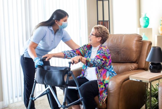 female caregiver helping a senior female out of a recliner with the help of a walker.  Caregiver is wearing gloves and a face mask