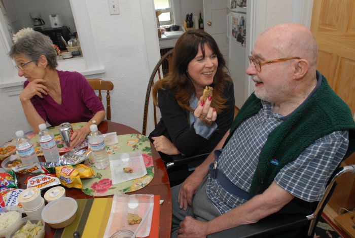 one male senior being helped at a dinner table by one female caregiver while another senior female is having a conversation off frame.