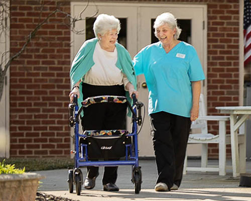 Female Caregiver Walking With Senior Woman Using A Walker At An Assisted Living Community