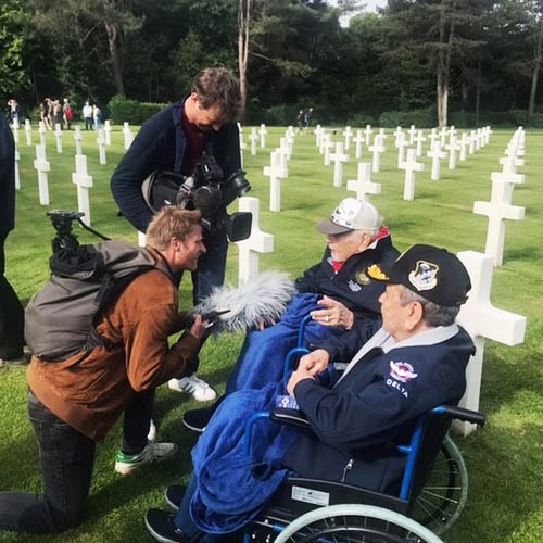 Senior Veterans in Wheelchairs in European Cemetery with Crosses in the Background and Photographers in the Foreground