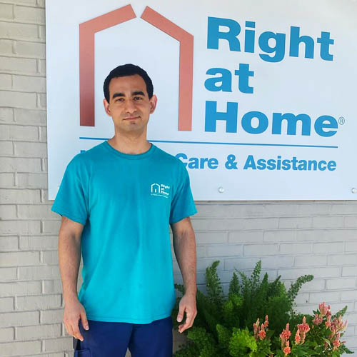 Young Male Right at Home Caregiver Posing Outside Next to a Right at Home Sign and Wearing a Right at Home Logoed Shirt