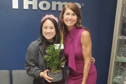 Two Women Pose for a Photo in Front of a Blue Background Holding a Plant at an Open House in Lafayette