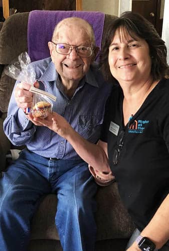 Female Caregiver Posing With Senior Veteran And Packaged Gift Muffins