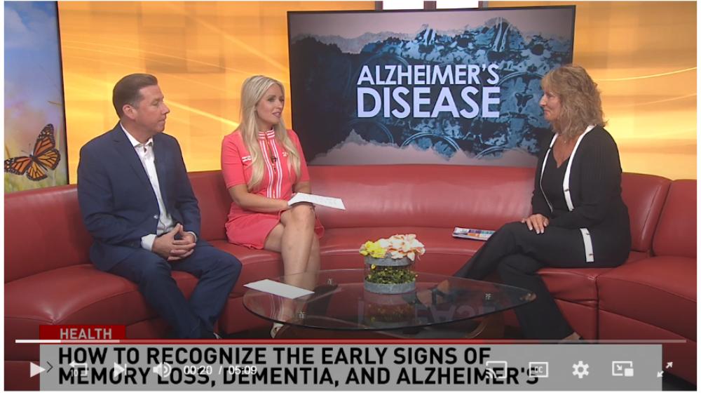 Three individuals are seated on a red couch in a television studio, engaged in a discussion. The person on the left is wearing a suit, the middle person is in a pink dress, and the person on the right is dressed in black. A glass coffee table with a floral arrangement and books is in front of them. An on-screen graphic reads “Alzheimer’s Disease” with an image of mountains and a butterfly. Below, another text overlay states “HEALTH - HOW TO RECOGNIZE THE EARLY SIGNS OF MEMORY LOSS, DEMENTIA, AND ALZHEIMER’S.” indicating a televised interview or discussion on Alzheimer’s disease and its early symptoms.