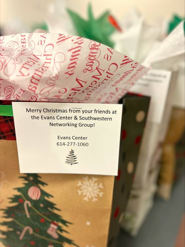 half of a holiday gift bag with a Christmas Tree on it with candy canes and balls with a note tag on it and printed tissue paper sticking out the top