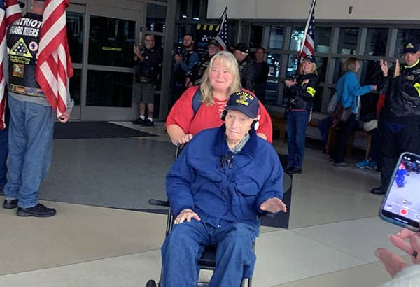 A woman pushes and elderly man in a wheelchair through an airport terminal. There are several people surrounding them, clapping. Two people are holding American flags.