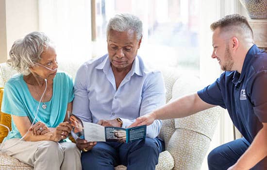 A senior man and woman sitting on a couch in a living room, reading a brochure while a Right at Home employee points out something in the brochure. 