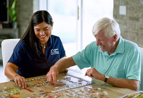 Female Right at Home caregiver helping a senior male put a puzzle together on a dining room table