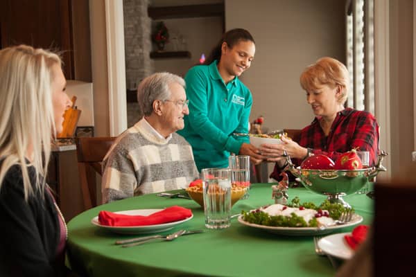 female caregiver helping serve a holiday meal to two seniors, one male, one female, and a younger female at a table in a dining room