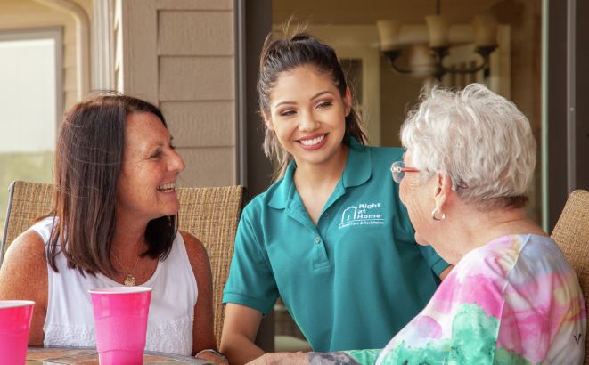 Female caregiver at table with senior female and her adult daughter while they are enjoying a beverage outside