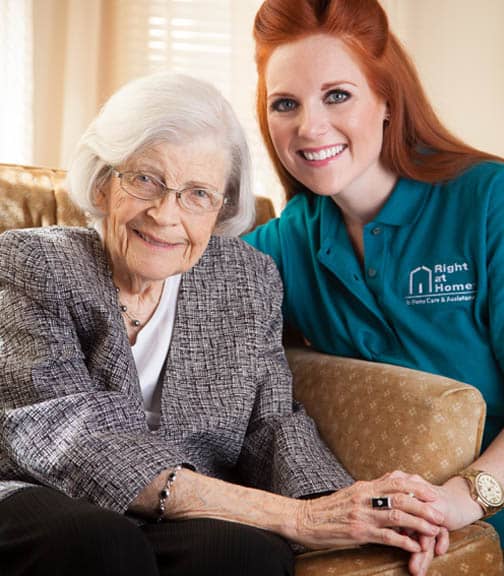 Senior woman sitting in a chair holding hands with a female caregiver with both smiling to the camera