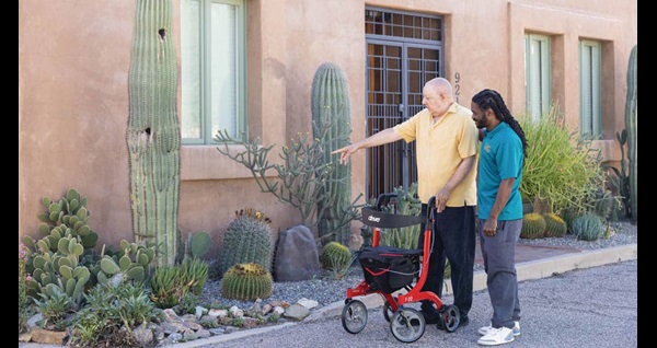 Male Right at Home caregiver standing next to male senior client using a walker outside looking at cacti on a sunny day. 