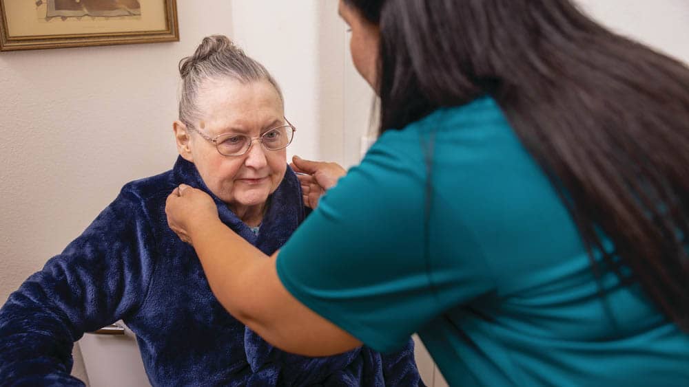 A senior female wearing a robe, getting assistance from a female right at home caregiver