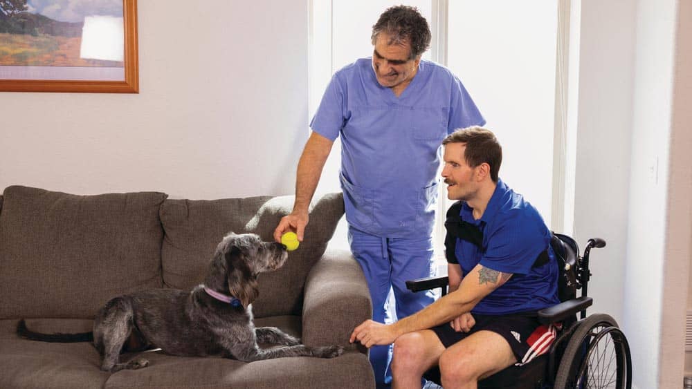 A disabled man in a wheelchair is next to his male Right at Home caregiver who is holding a tennis ball up to a dog laying on the couch. 