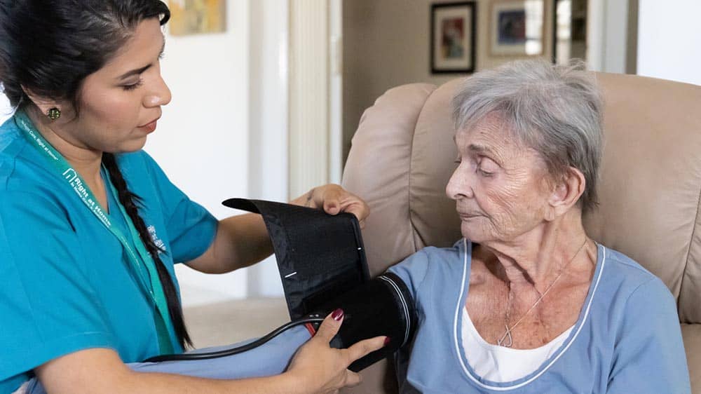 A female Right at Home caregiver is applying a blood pressure cuff to a senior female client's arm while the client is sitting in a recliner