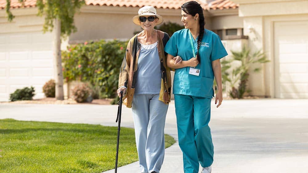 Female Right at Home caregiver assisting a female senior client, using a cane, walk down the driveway