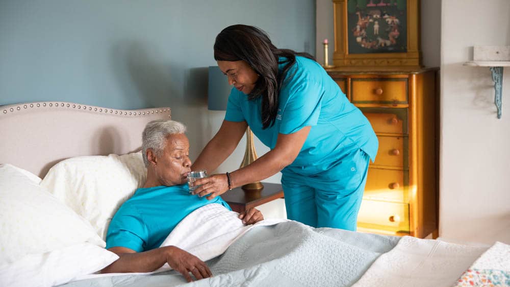 Female Right at Home caregiver helping a senior male client take a drink of water while he is laying in bed