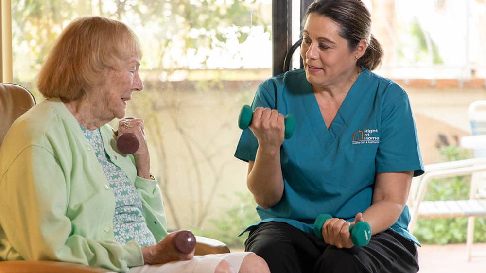 Female Right at Home caregiver helping a senior female do arm curls with small weights