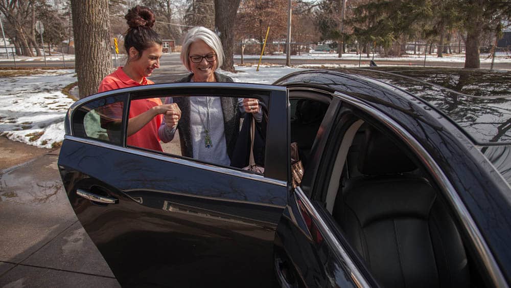 Female Right at Home caregiver is helping a senior female client into the backseat of a car