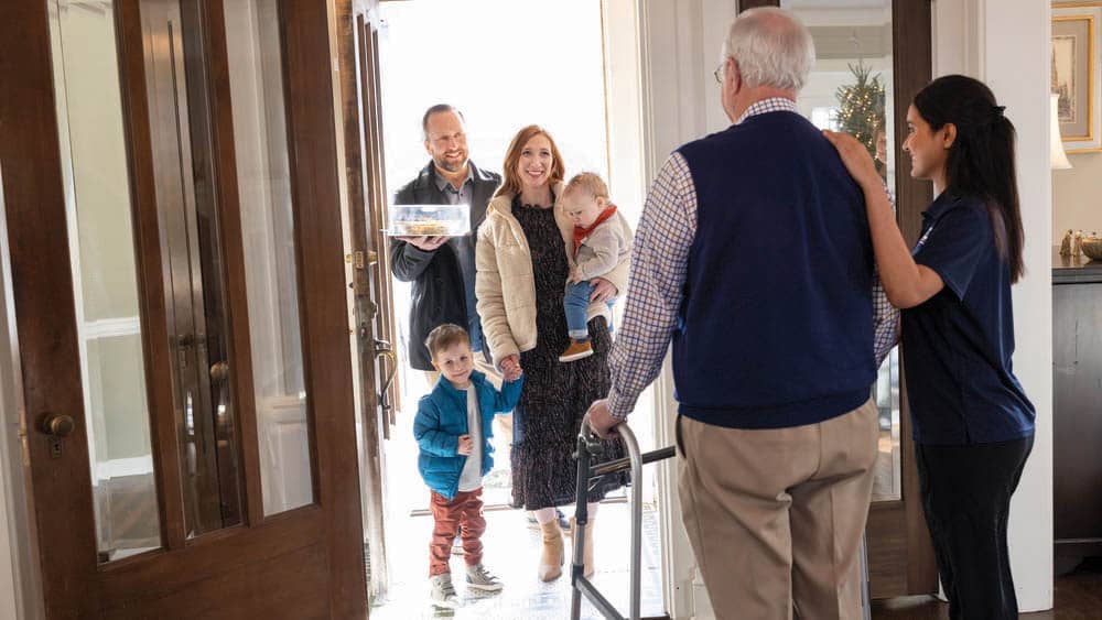 A senior male client, along with his female Right at Home caregiver, greet members of the senior's family at the door.