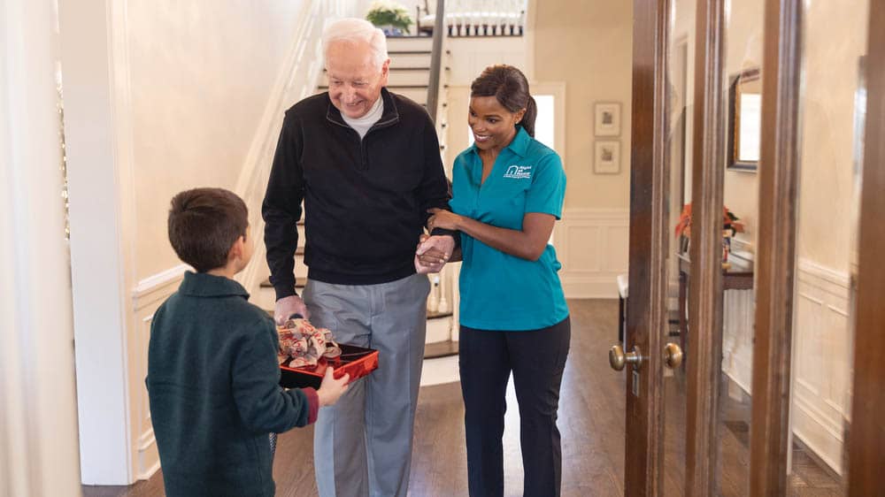 A young boy is handing a senior male a Christmas present in the entryway of a home, while a female Right at Home caregiver is helping the senior male stand 