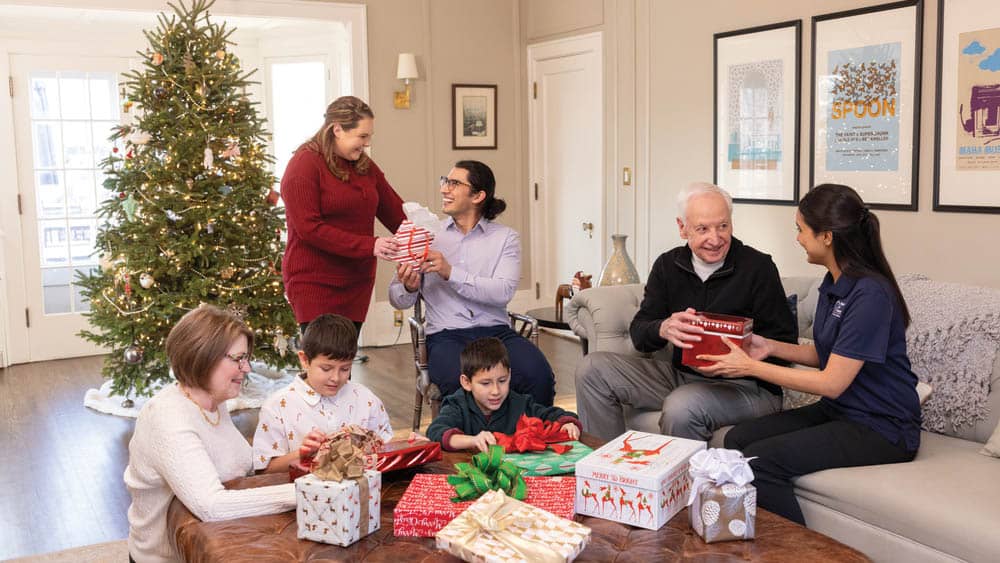 A multigenerational family consisting of two senior grand parents, two parents, and two children opening Christmas gifts in a living room with a large Christmas tree, while a female Right at Home caregiver joins them.